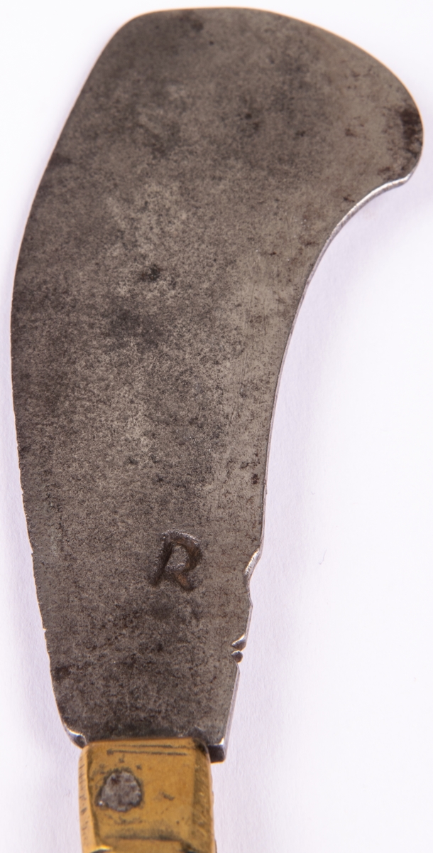 Gästabudskniv med brett knivblad att fälla in i mässingskaftet. Gaffel, tvåhornig av järn. Hållare av mässing, skaft av mässing.
Skaftet har dekoren av ett drakhuvud.
"R" stansat i bladet.