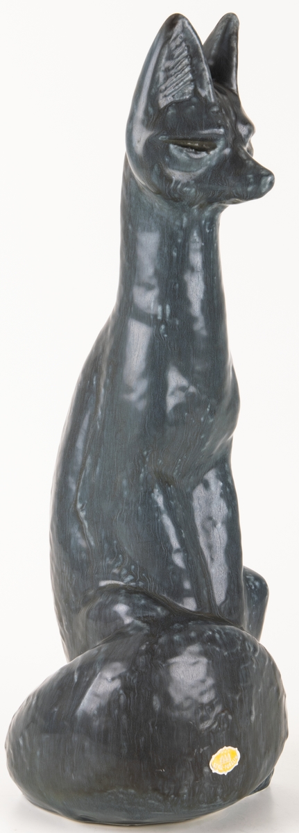 Figurinen Räv av formpressad flintgods, formgiven av konstnär Lillemor Mannerheim. Grågblå glasyr. Sittande räv med svansen lindad runt nedre delen av kroppen.