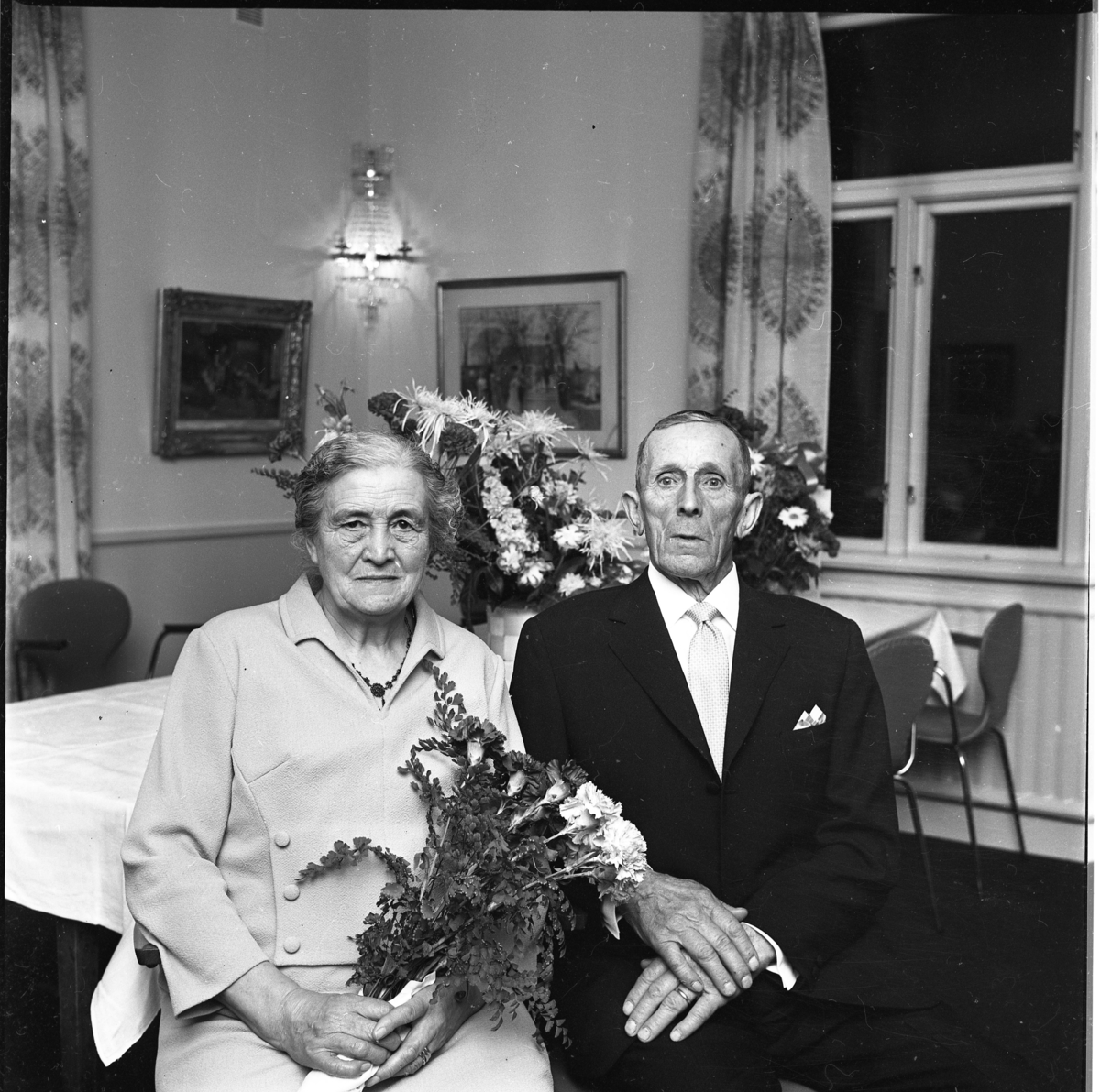 Ett äldre par sitter i sitt hem, kvinnan har en blomsterbukett i handen. Det är Elsa Johansson och maken Gunnar Pettersson som firar guldbröllop.