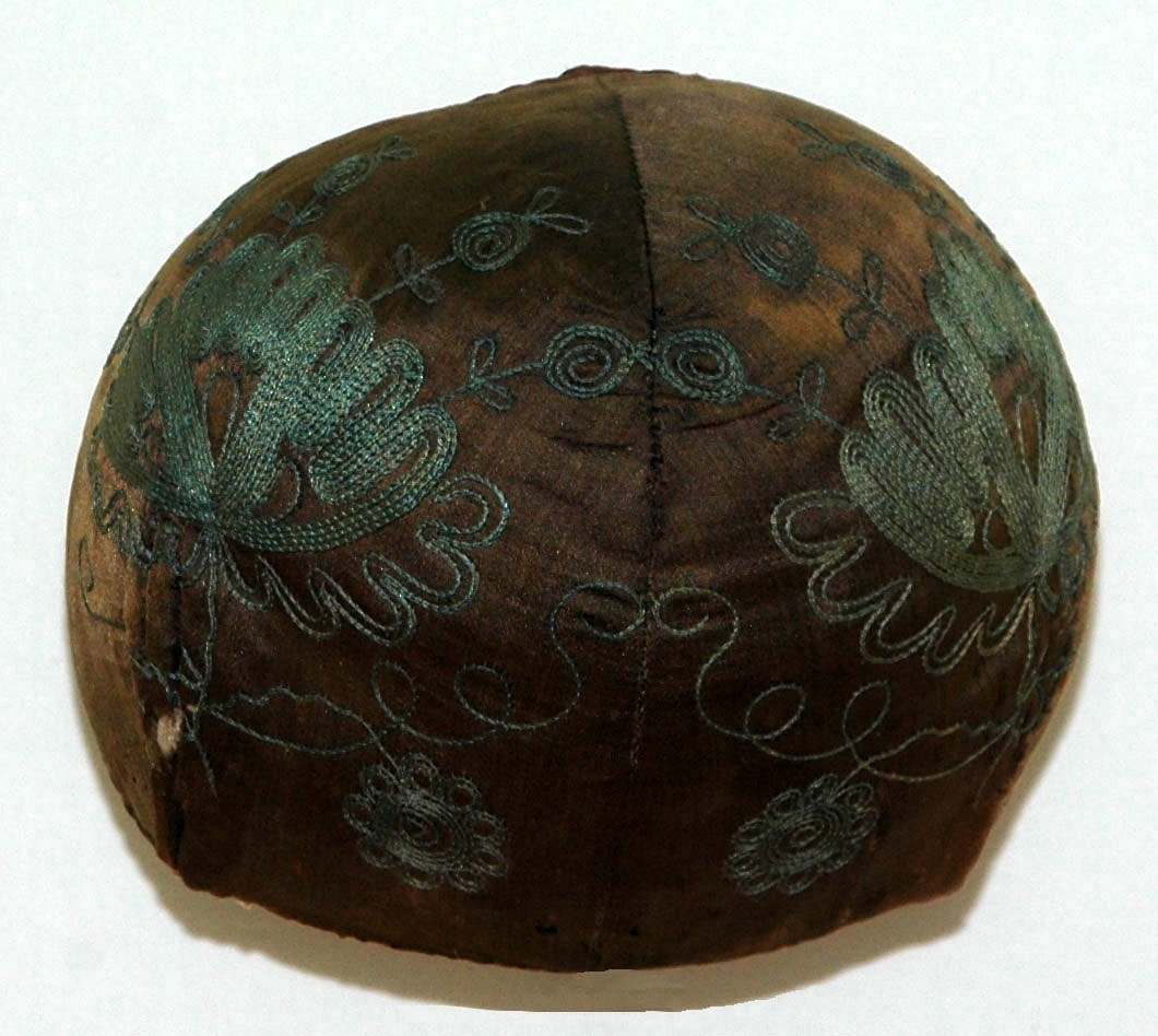 Bindmössa av brun-svart sidensatin försedd med tambursömsboderi i blått. Fodrad med naturfärgad linnelärft.