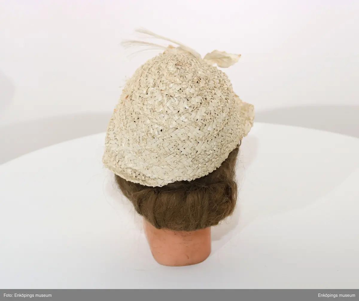 Krämvit hatt tillverkad av spiralsydda bastband. Brätte på halva rundeln. Dekor av två blomknoppslika former sydda av vit sammet, även stjälkar och blad av textil. Två " tofsar av tagel". Tillverkaren heter Ingrid Wik, Enköping, brukaren till denna hatt var Britt Lundevall.