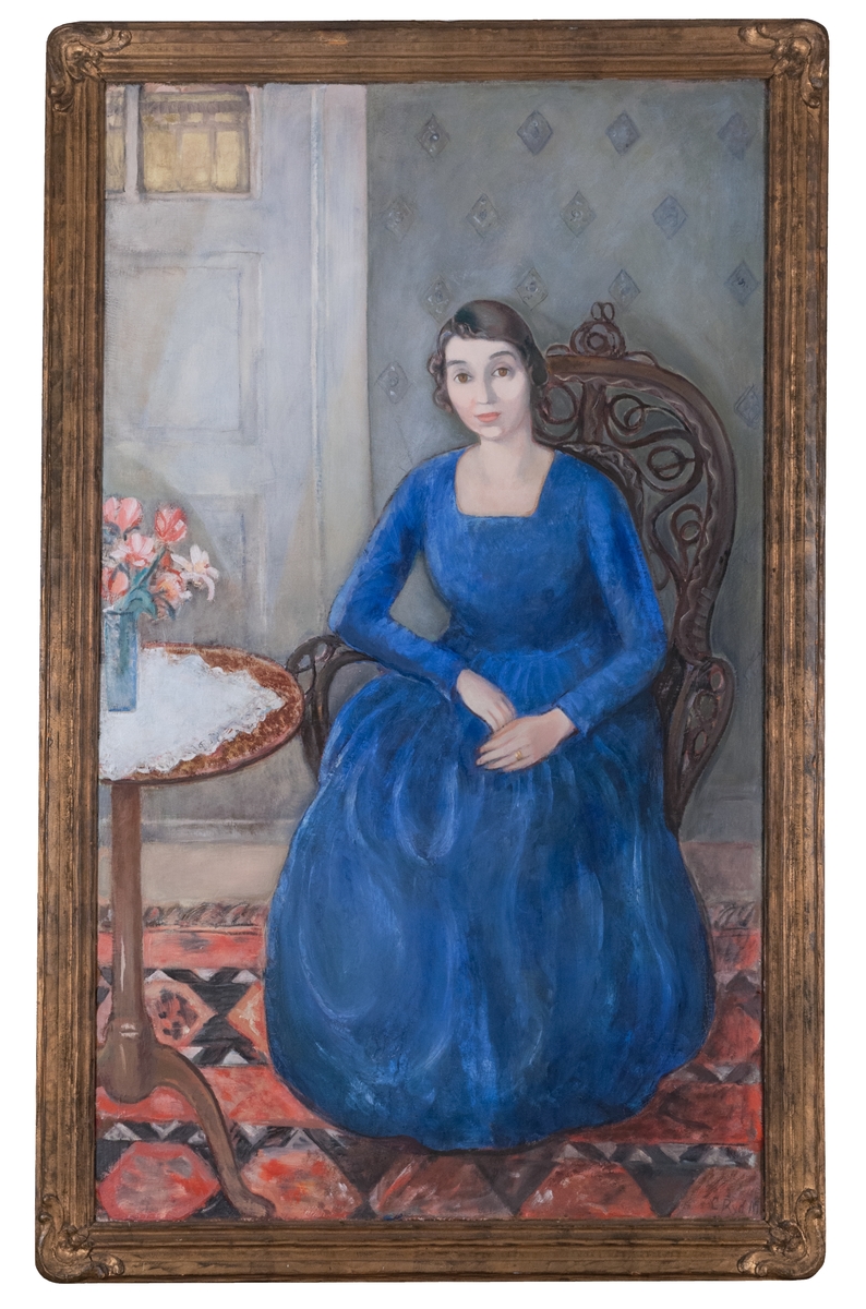 Oljemålning på duk, "Konstnärens hustru" av Carl Ryd. Kvinna i helfigur sittande på rottingstol vid litet, runt sybord; hon bär blå klänning, på bordet vit spetsduk och smal glasvas med skära och vita blommor; i fonden grå tapet samt dörr.