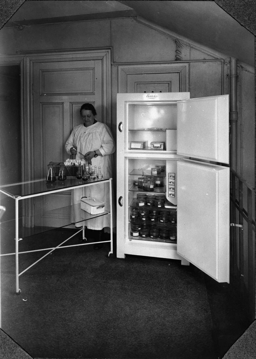 Electrolux kylskåp typ D i "Statsmedecinska Anstaltens Bakteriologiska Avdelning". Kylskåpet användes för förvaring av vaccin. Genom kontinuerlig drift erhålles i nedre avd. en temperatur av -2 grader Celsius. >> Kvinnan hämtar vaccin??