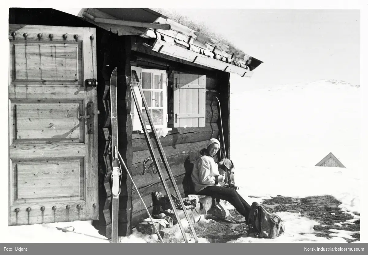 Kvinne sitter langs husvegg ved hytte. Ski står langs husvegg