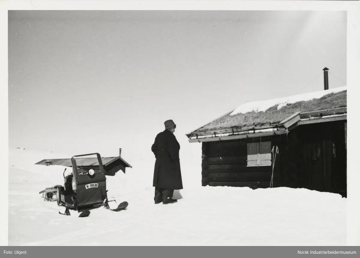 James Coward står i snøen ved snøskuter og hytte med torvtak og lukket vinduslem