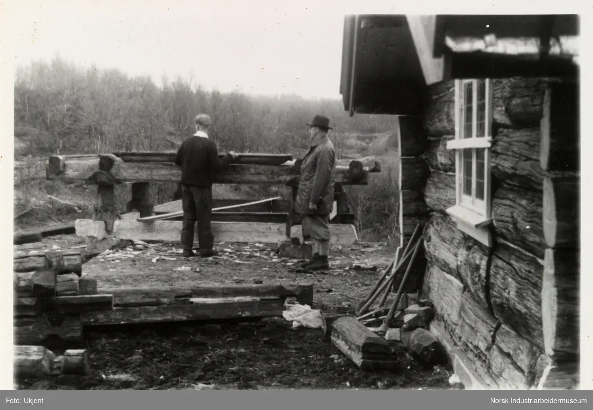 To menn står ved grunnstokker til bur på Sundet. Til høyer sees laftet hytte og redskaper