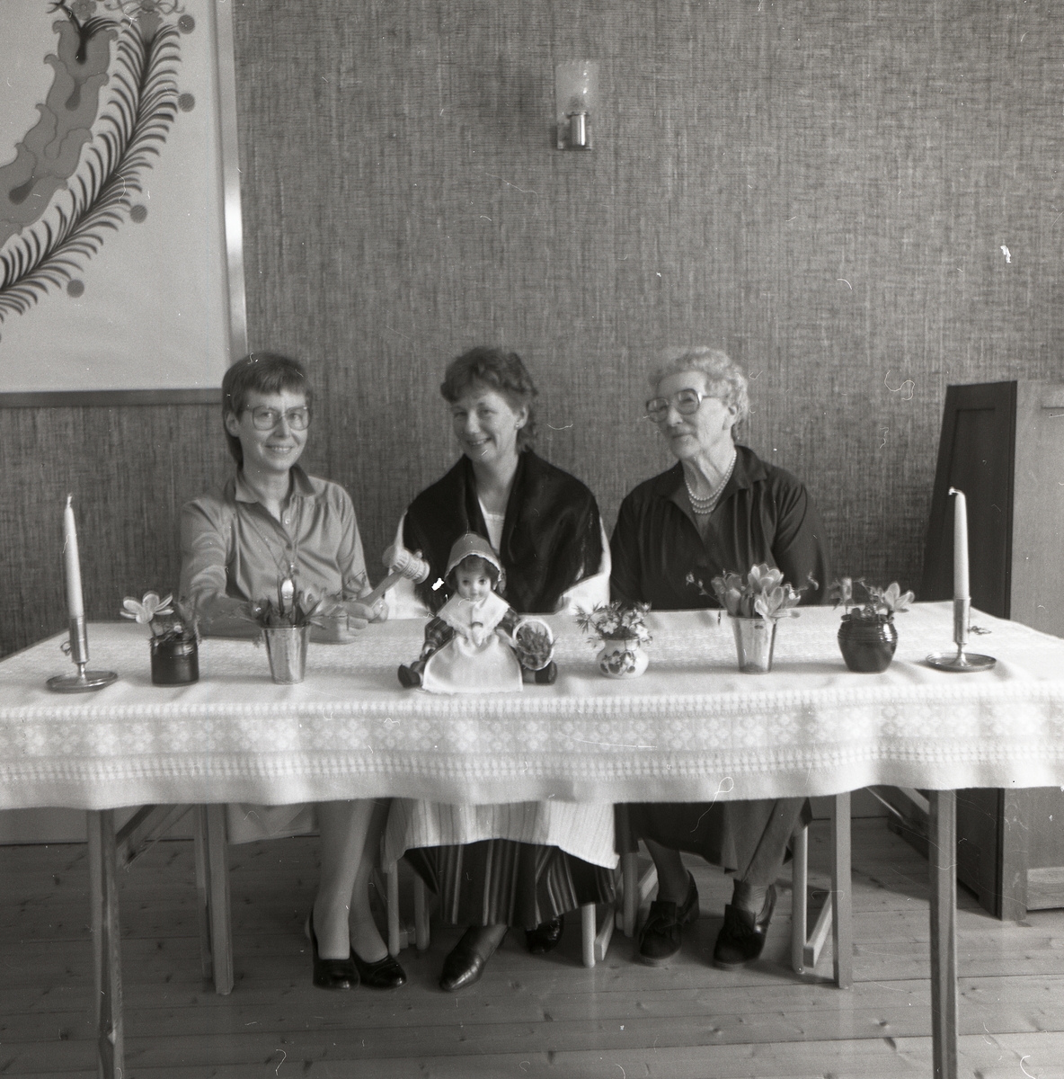 Tre kvinnor sitter vid ett bord dekorerat med vaser, ljusstakar och en docka under husmodersföreningens länsårsmöte, 26 april 1986.