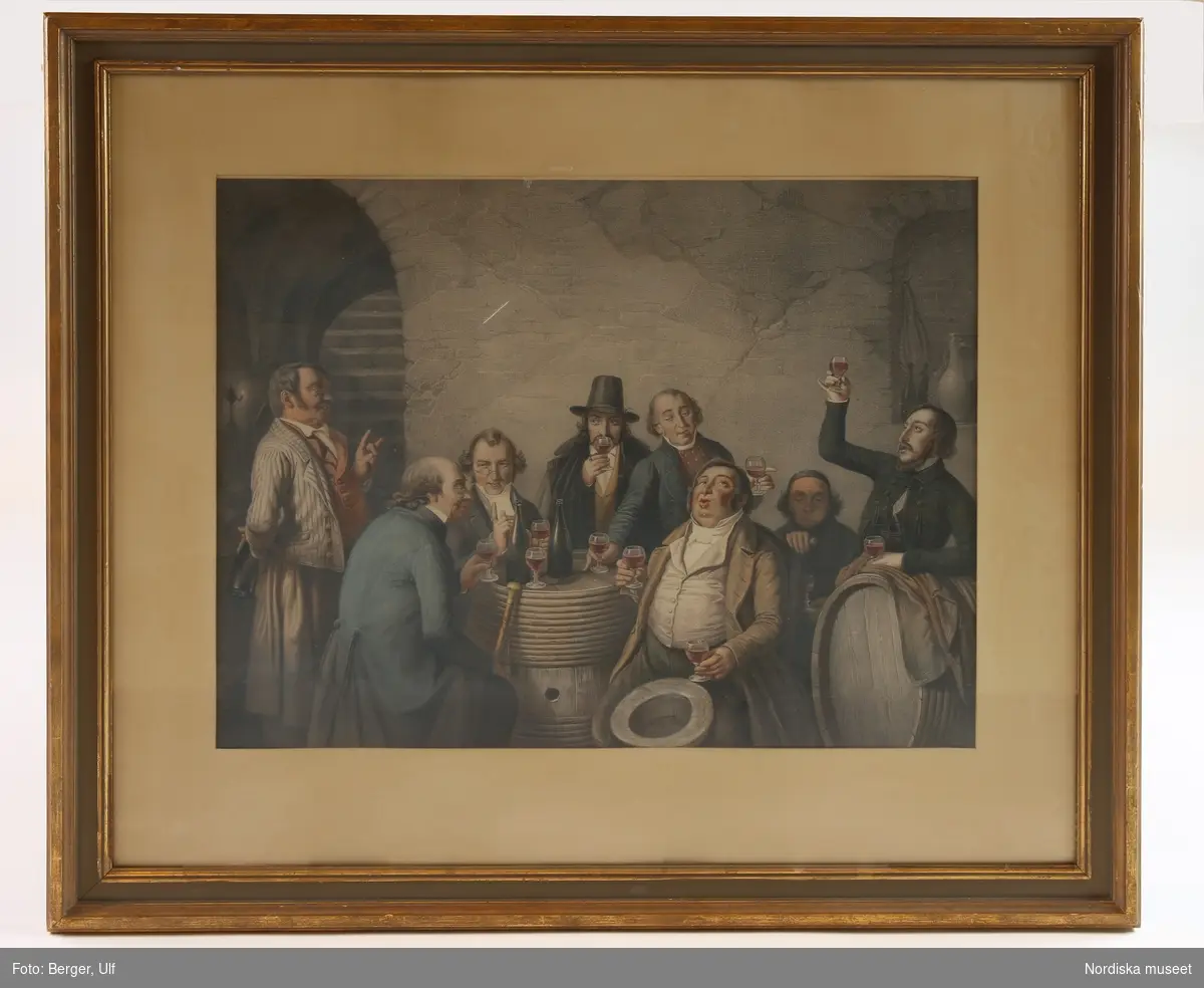 En grupp män dricker rött vin ur glas samlade kring vinfat i en källare.