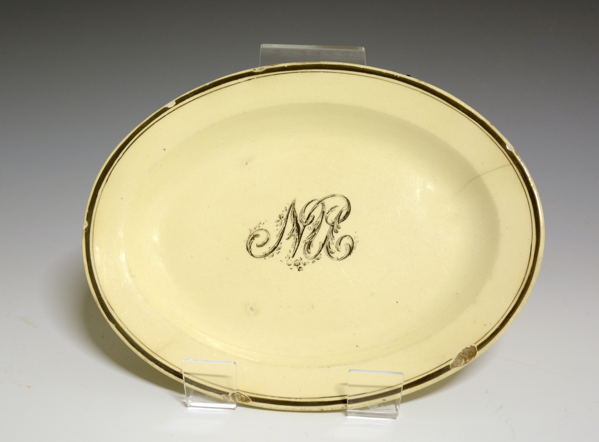 Lite, ovalt fat av stentøy med gulhvit glasur. I speilen et monogram med bokstavene NU. Ytterst på fanen en bredere og en smalere sort strek.