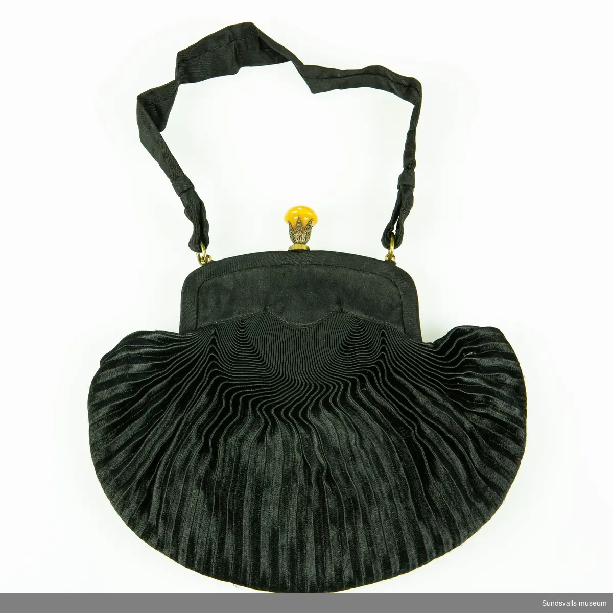 Aftonväska i svart plisserat tyg med bärnstensfärgad knopp. Inuti en på rem fastsatt spegel, samt fastsatt börs.