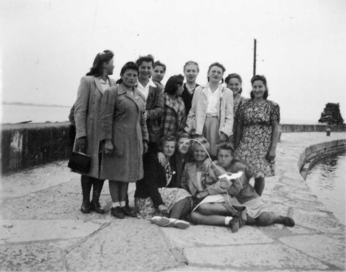 Tretton polskor stående och sittande på en hamnpir, troligen i Gränna. Några har kappa eller jacka på sig. De skrattar och ser glada ut.