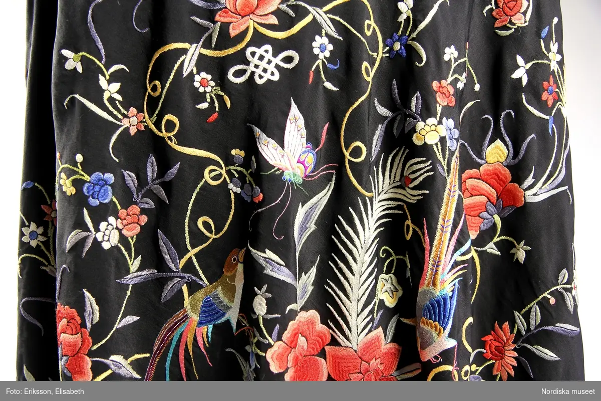 Kvadratiskt formad schal av svart sidentyg med flerfärgat broderi med motiv av växter och fåglar. Långa, svarta fransar (37 cm. långa) längs sidorna.  
/Lisa Eide 2015-06-16