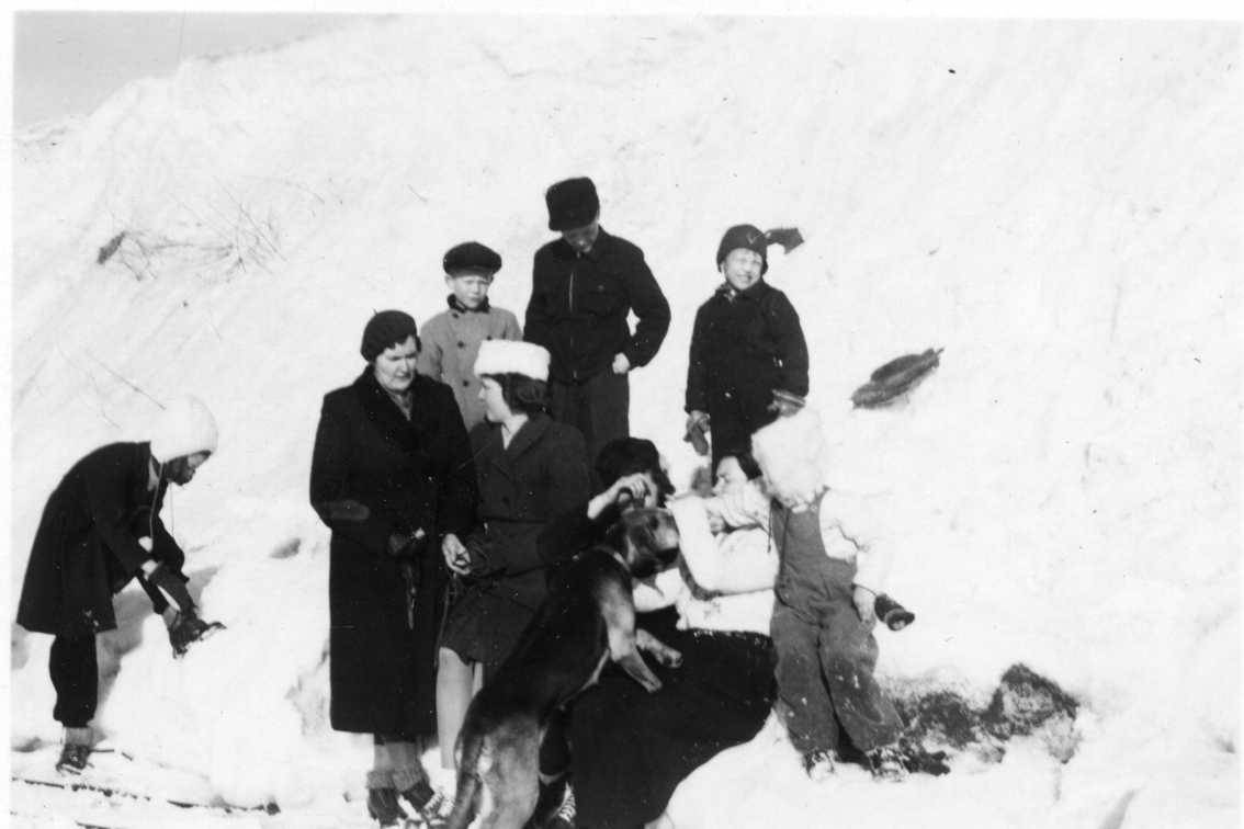 Vuxna, barn och en hund står och sitter i motljus vid en hög snötäckt vall. På marken ligger möjligen skidor och någon av dem kan ha skridskor på fötterna. En av kvinnorna heter Ingeborg Idh.