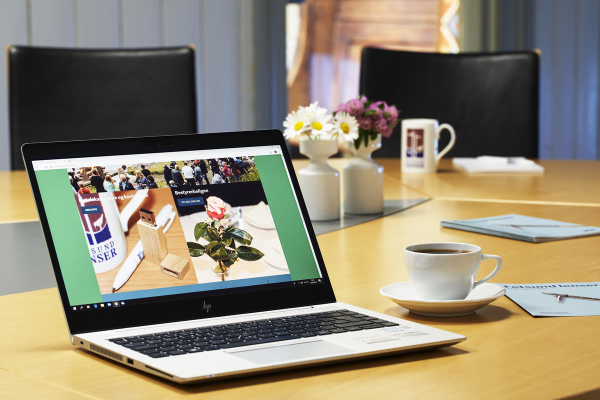 Bærbar pc i forgrunnen med kaffekopp og folder på høyre side på et konferansebord. Blomster og kaffekopper i bakgrunnen