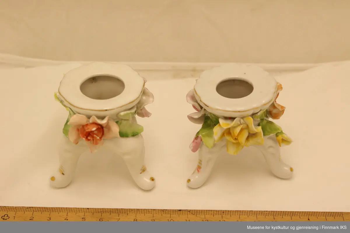 To små lysestaker i porselen. Lysestakene står på tre bein, og har dekorative blomster og blader rundt kanten, og med gullfarve malt rundt kanten og på hvert av bena.