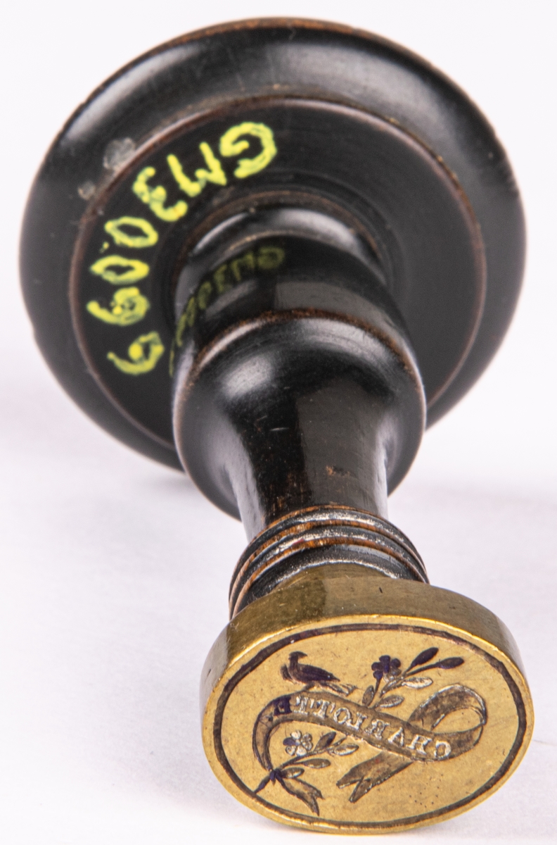 Sigillstamp i mässing med svarvat trähandtag. Stampen är oval med fågel (duva), blomstängel och textband med namnet "CHARLOTTE".