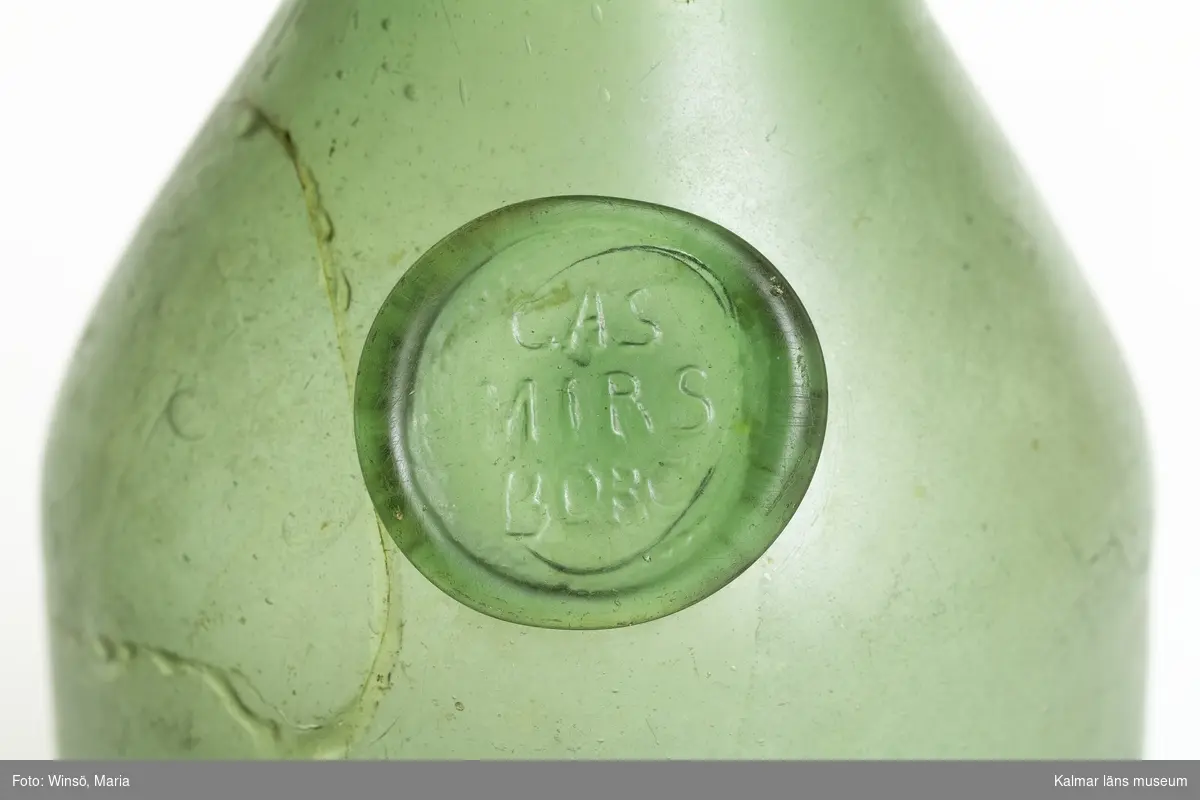 KLM 46239. Flaska. Av glas. Cylindrisk, gröntonad glasmassa. Sigillmärkt: CASMIRSBORG. Skada på skuldran, lagning, en bit har limmats på plats samt längre spricka. Förvärvad i detta skick.