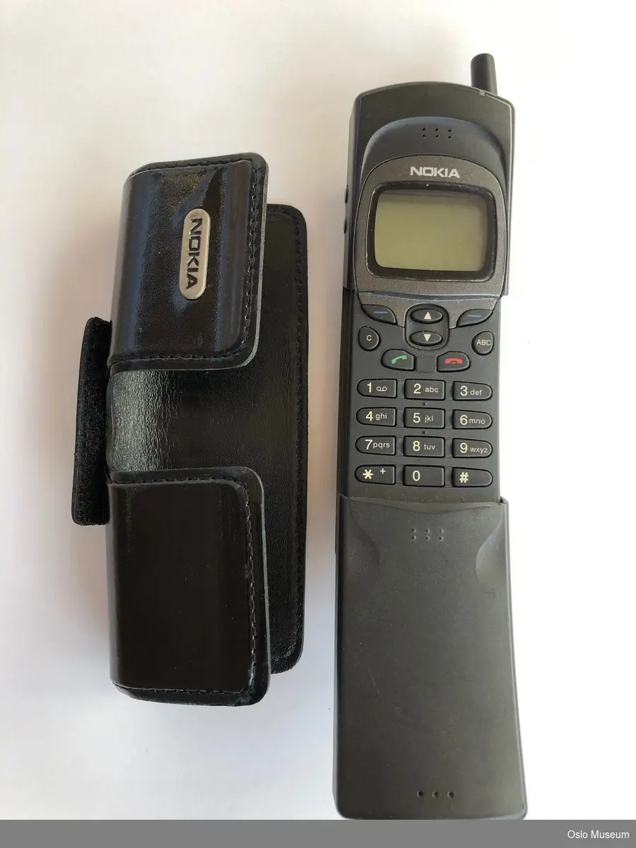 Mobiltelefon i plastmateriale med nedfellbart tastelokk, utenpåliggende gummitaster og fast antenne. Taske i skinn med borrelås til å feste i beltet.