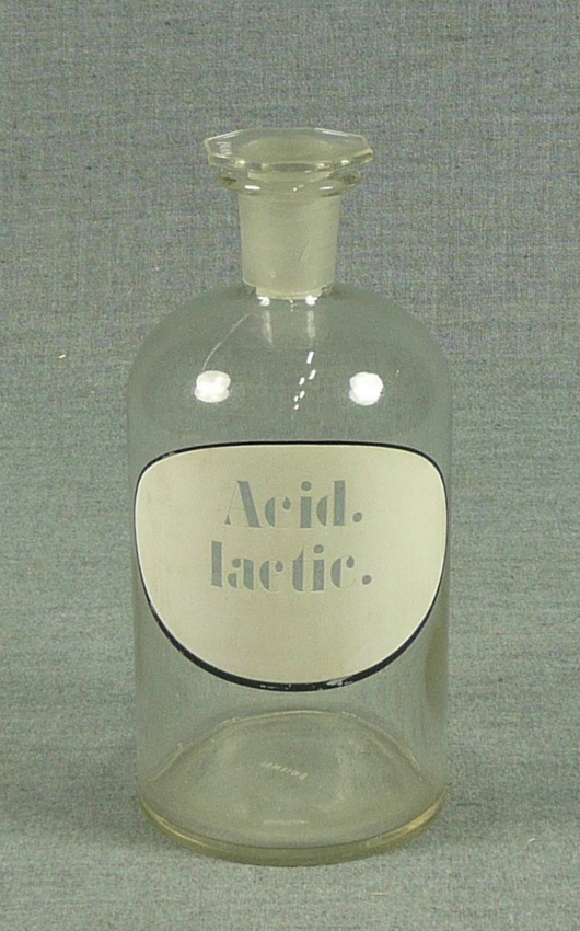 Drogkärl av transparent glas med smal hals och oktogenal glaspropp. Vit etikett med texten Acid Lactic.