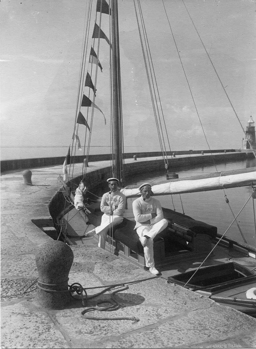 Två unga män med armarna i kors och pipa i munnen sitter i von Dübens segelbåt Blenda i Gränna hamn. De bär ljusa byxor, stickad ljus tröja med broderat "BLENDA" på bröstet och sjömansmössa på huvudet. De väntar på att föra kronprinsparet Gustav Adolf och Margaret tillsammans med herr och fru von Düben till Visingsö. Mannen till vänster är Carl Wahlberg och till höger sitter Emil Carlsson.
