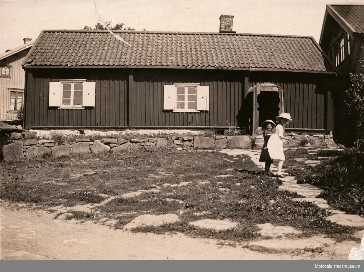 Porträtt av två flickor framför det troligen äldsta huset i Fässbergs by. Fässberg Västergård 6, Fässberg 1:34. Fotografiet är taget ca 1910. Flickan i den stora hatten är fotografiets ägares farmor Edit Olsson, född 1903 (gift 1923 Andersson).