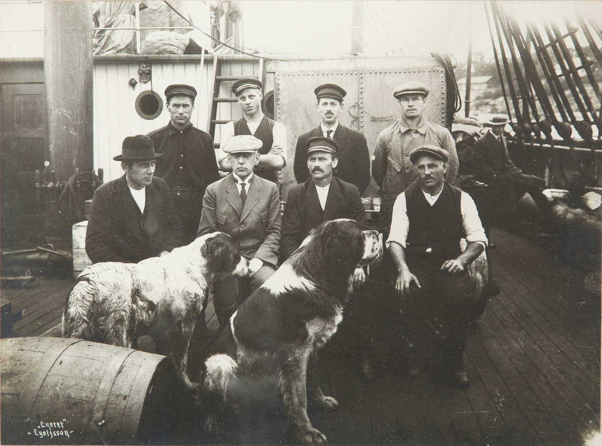 Gruppebilde av Roald Amundsen og (antall?) menn på båtdekk, med 2 hunder