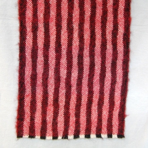 Randig halsduk vävd i tuskaft av ullgarn. Varpen är randig i svart och naturvitt, inslaget är i två röda nyanser i 125 mm breda partier.Halsduken är formgiven av Ann-Mari Nilsson.