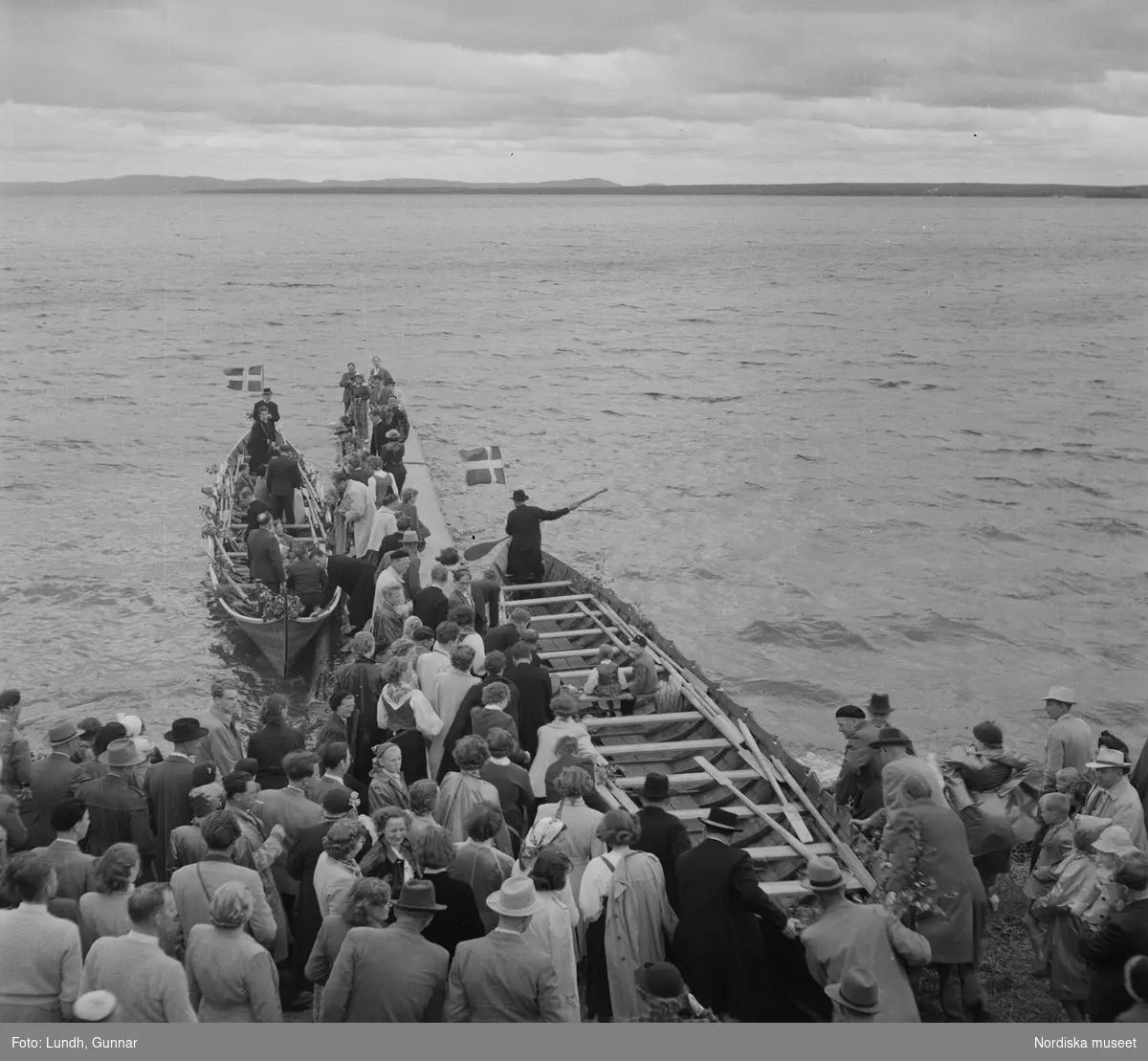 Motiv: (ingen anteckning) ;
En folksamling vid Rättviks kyrka, en folksamling står vid en brygga när kvinnor och män går ombord på två kyrkbåtar.