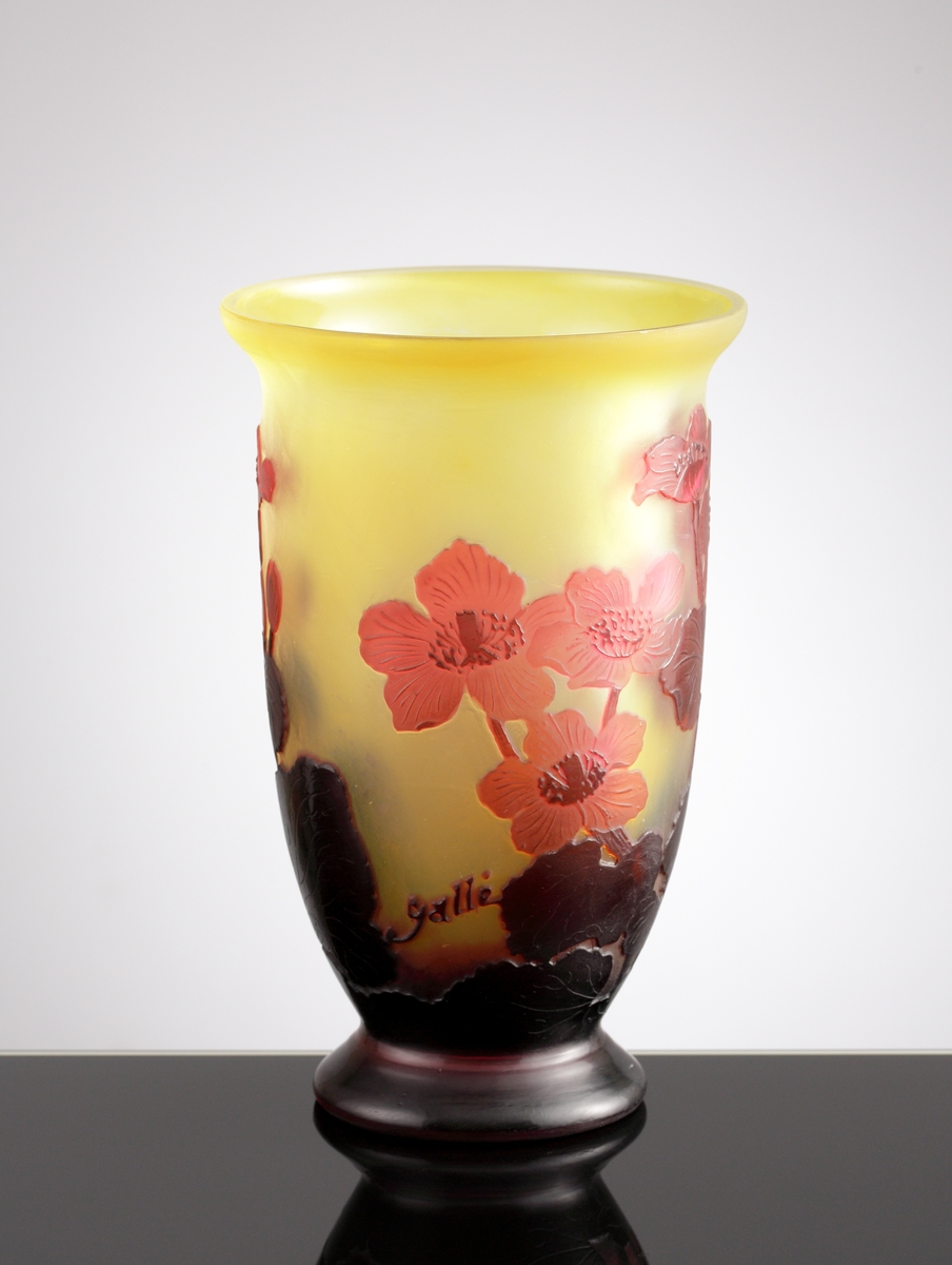 Halvopak, gulgrön vas med etsat överfång i röda nyanser som avslutas i en mörk fot. Växtmotiv med blommor och blad.