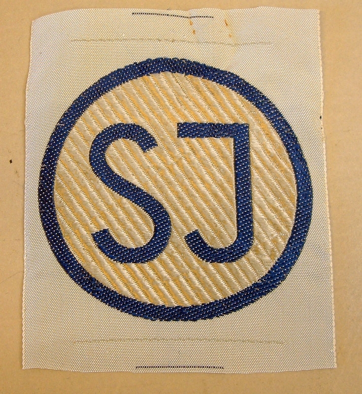 SJ tygmärke, med botten i silverfärgat randigt tyg. Rund ring med SJ i mitten, i blått.