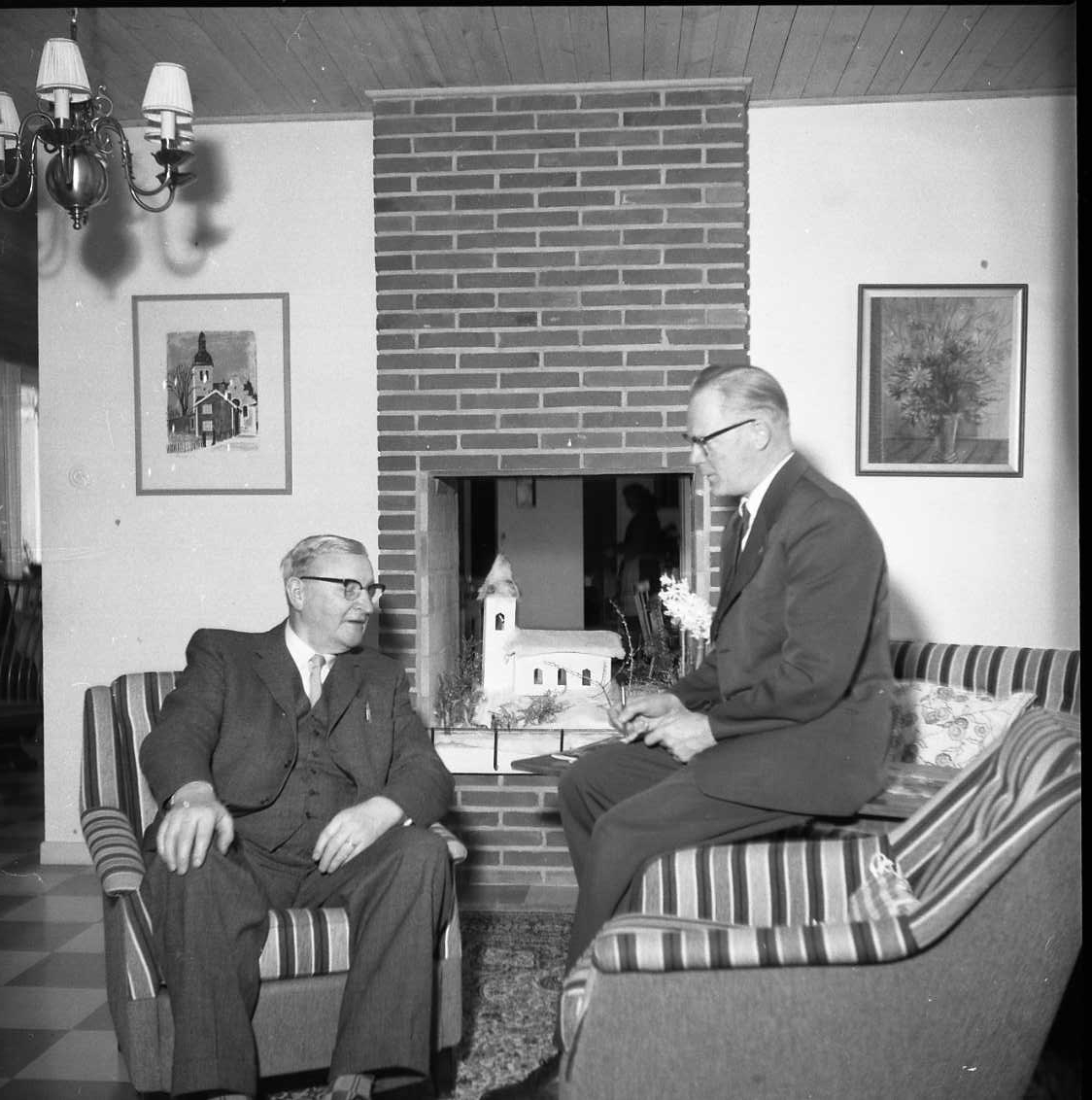 Interiör från en hemmiljö. Gustaf Rahm sitter i en fåtölj och samtalar med Percy Heldebrandt som sitter på ett bord.