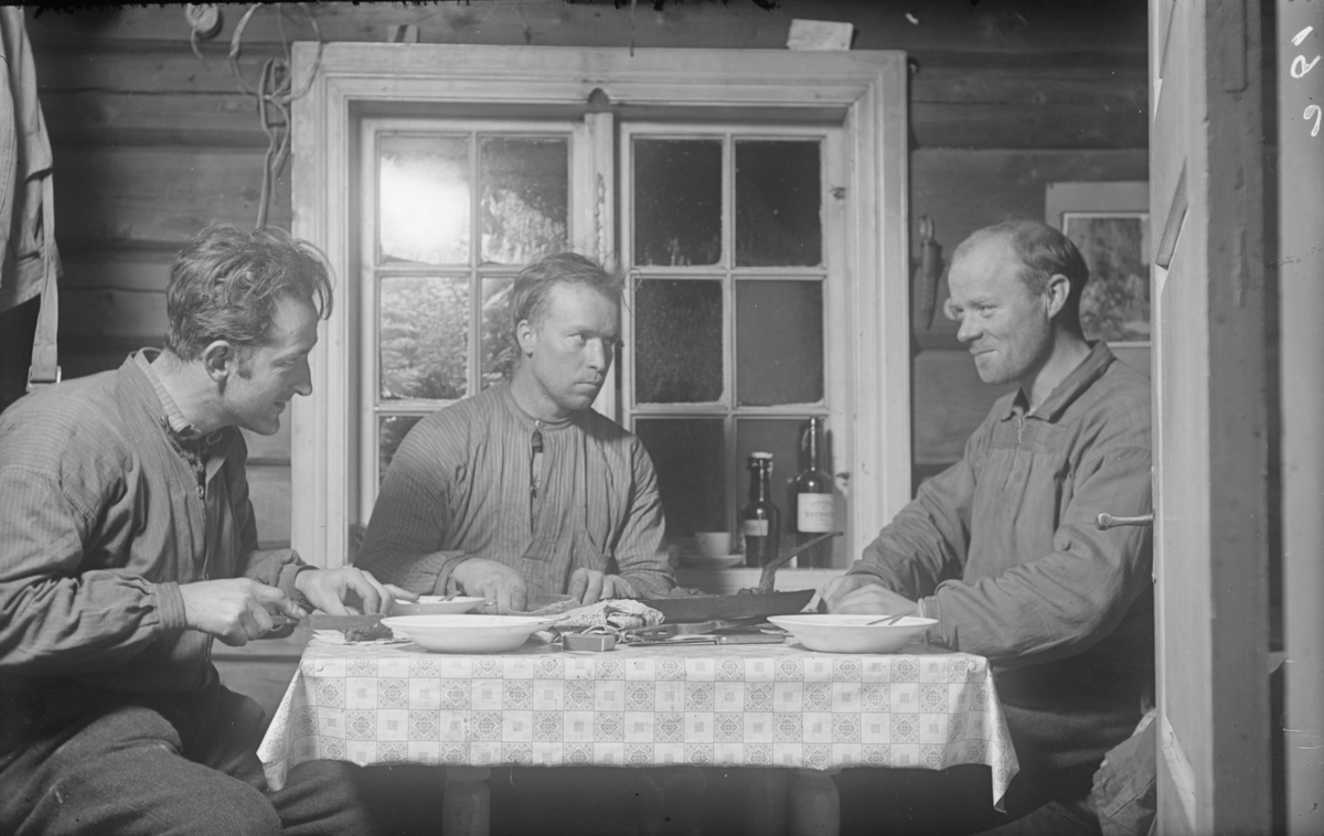 Tre menn har et måltid i ei tømmerhytte. Til venstre fotograf Karl Olav Moe.