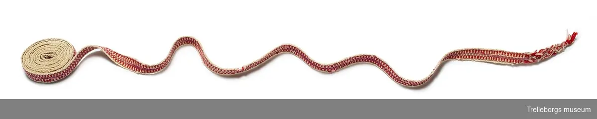 Vävt ullstrumpeband i vitt, rött, vinrött och lila. Ena änden är tvärt avklippt, den andra är flätad ca 8 cm och därefter ihopknuten.