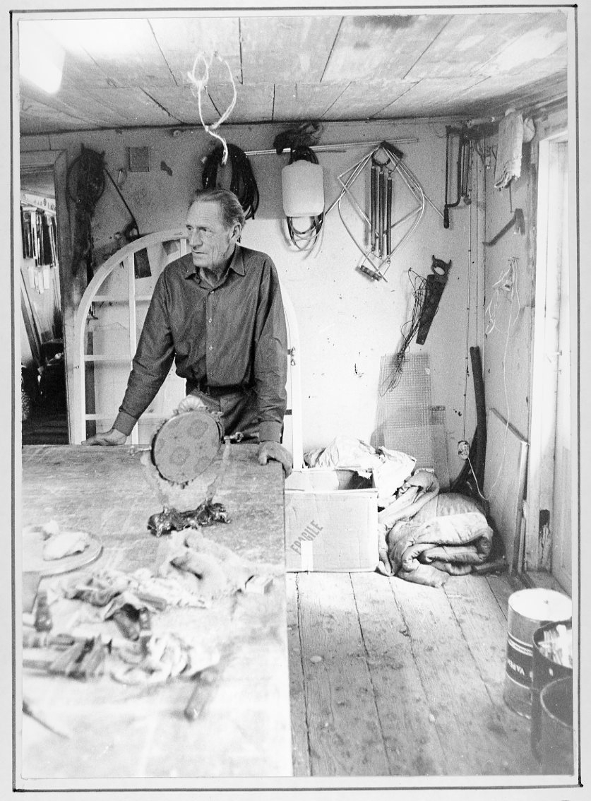 Porträtt av glasmästare Bernt Rendahl i glasmästeriverkstaden. Han står lutad mot ett bord. Mot väggen bakom honom står det en stor välvd fönsterbåge med spröjs. På väggen hänger det olika verktyg.
