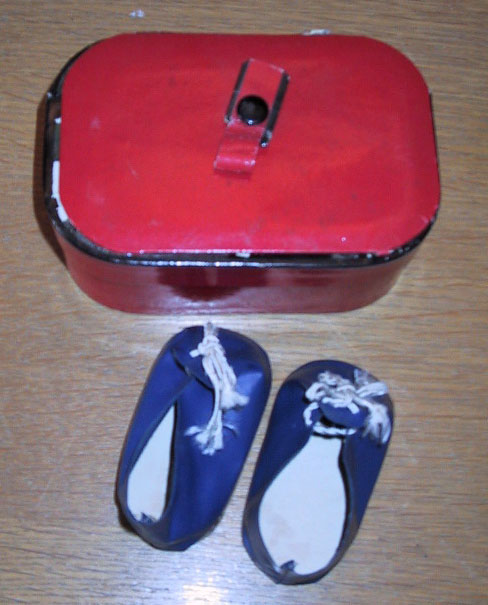 Rød koffert med trykknapp og svart kant, og et par blå dukkesko med hvite lisser.