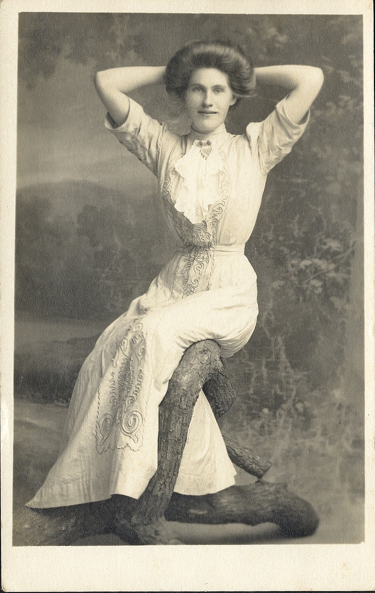 En okänd kvinna i ljus, broderad klänning med hög krage. Vid kragen syns en brosch. Hon sitter på en trädgren. 
Fotot är tryckt som vykort, möjligen i USA.