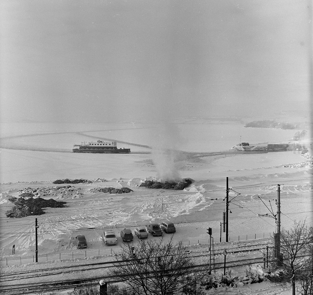 Hamar-Kapp-Ferjen, mjøsbåt, i råk i mjøsisen, utenfor Hamar Brygge, senvinteren 1967. Bål, avfallsbrenning, bygningsrester åttemetersplanet. Fergetrafikk.