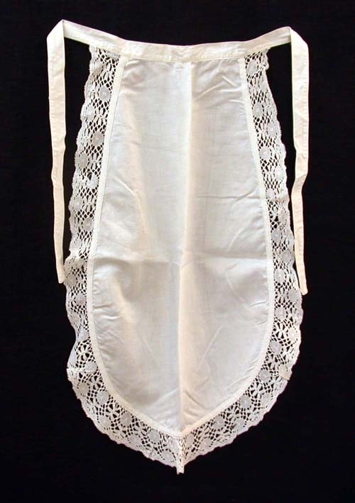 Ovalt serveringsförkläde i vitt, satinvävt bomullstyg, kantad med ca 50 mm bred spets. Cirka 20 mm bred linning som går i ett med knytbanden.