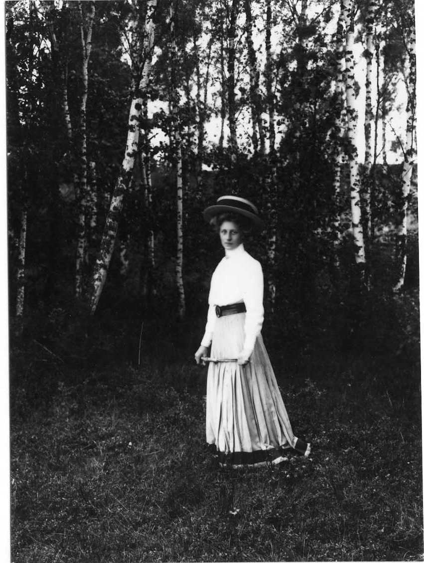 Porträtt av Ellen Gustavsson från Örserum. Hon står i en skogsglänta i lång smalrandig och plisserad kjol med mörk bård i nederkant. Ljus blus och bredbrättad ljus hatt med ett mörkt band.