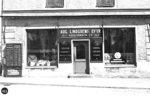 Magasinet, som uppfördes av korsvirke 1862 (brf), har bl a. inrymt en "Förvaringsbod för Mekanisk Brandstege" 1917-1928 och sistnämnda år inretts till två butiker i bottenvåningen. (Stig Pärson)  Butiken på bilden är AUG. LINDGRENs EFTR.  KRISTENSON & Co.