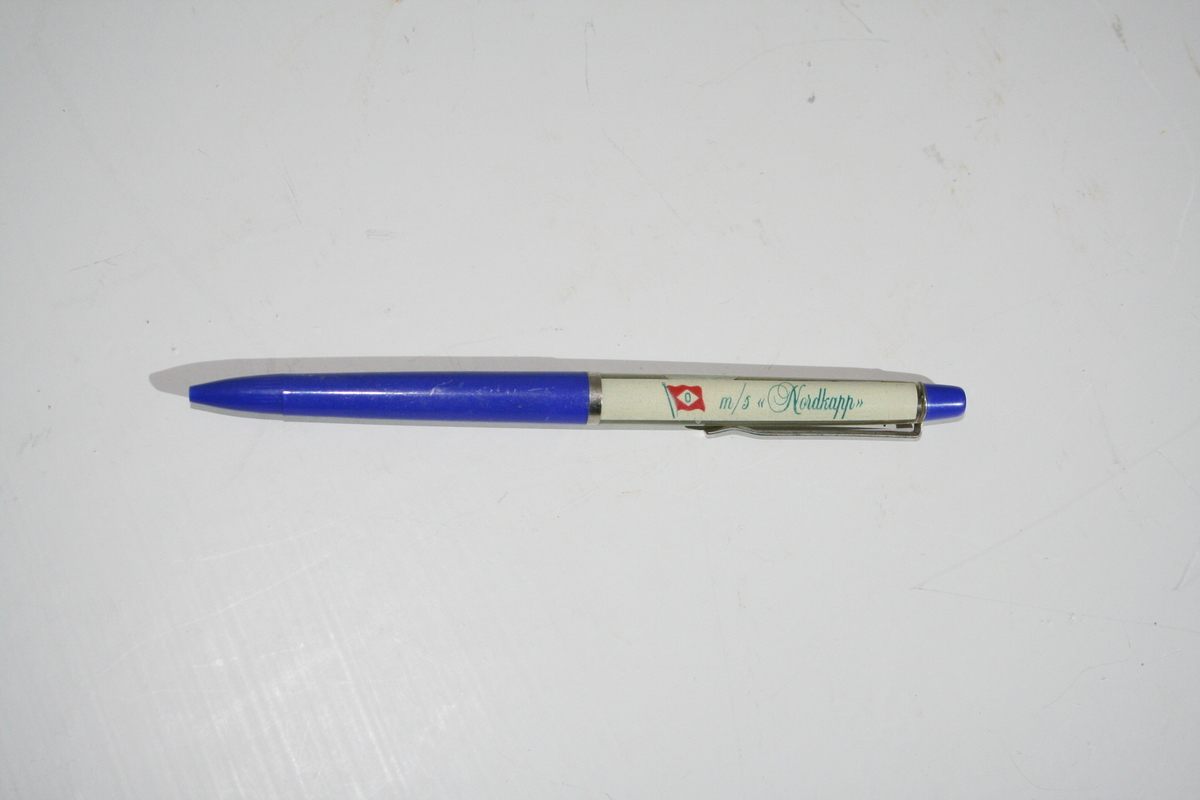Plastikk kulepenn. Suvenir fra M/S "Nordkapp". Pennen har en "flytende " hurtigrute i skaftet som passerer Nordkapplatået. Rederiflagget til Ofotens dampskibsselskap er tegnet på.