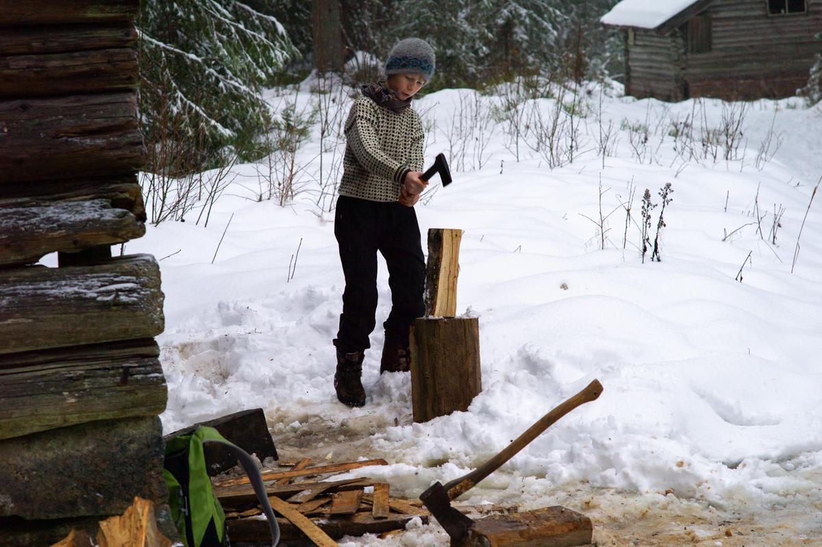 En yngre gutt med strikkegenser står og hogger ved ute mellom to tømmerbygg på Odalstunet. Det er vinter.