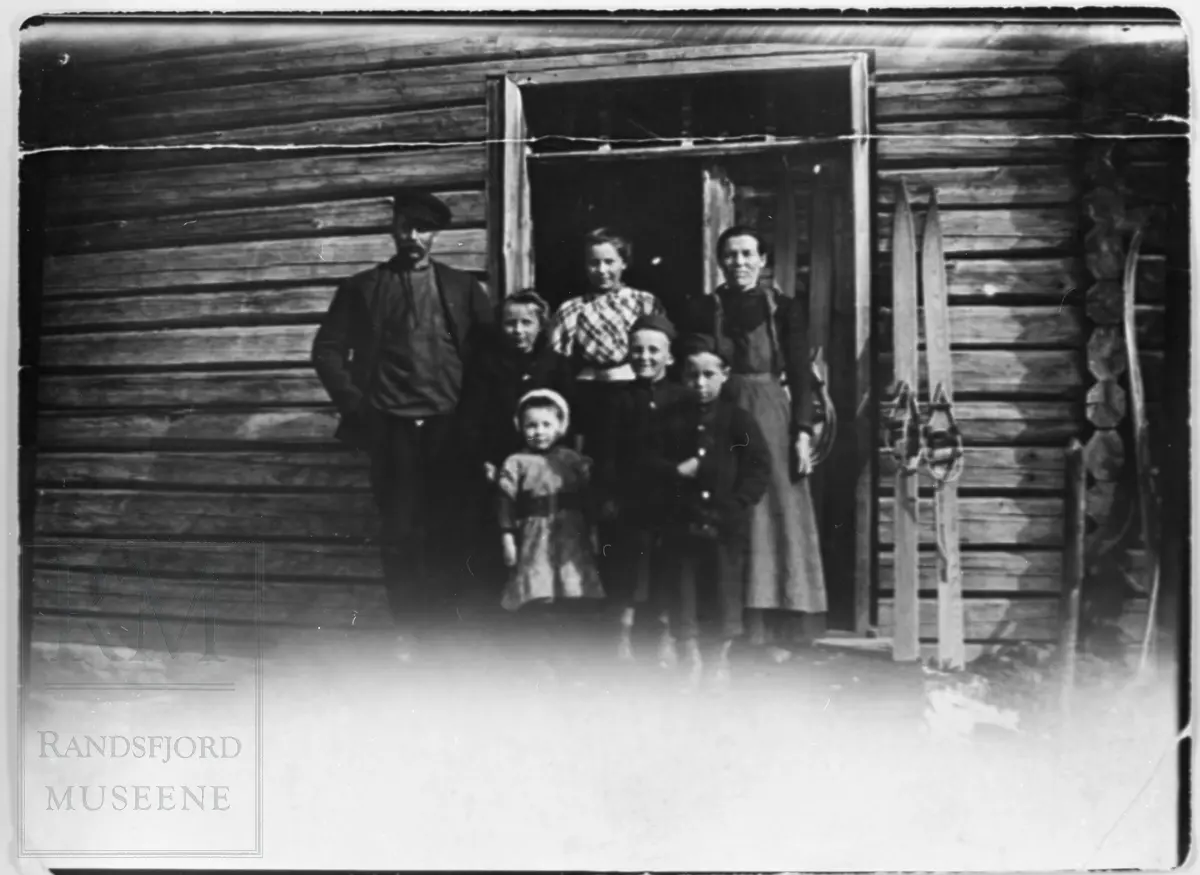 Gruppe av 4 barn og 3 voksne står oppstilt ved ei dør i en tømmerbyggning, familien på Våja ca 1912.