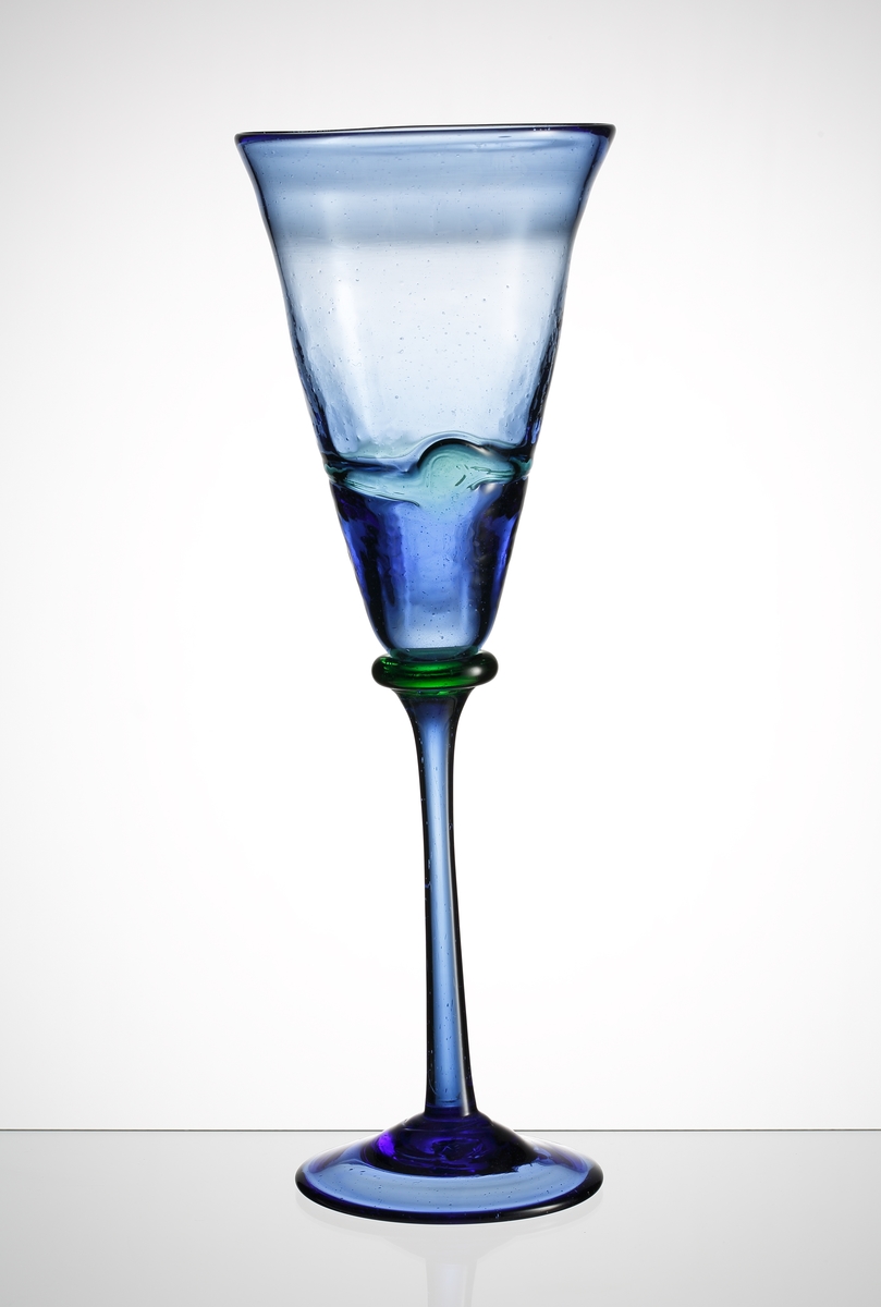 Blått högt vinglas med en blå, något kraftigare, pålagd tråd på kupans nedre tredjedel.
Glaset är blåst i återvinningsglas i paret Klenells hytta i Edsbjörke, Värmland.