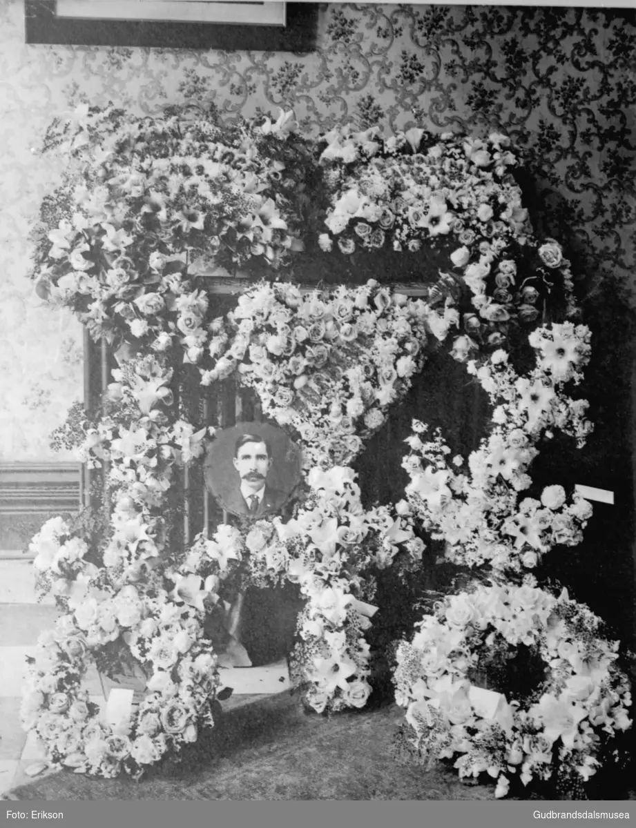 Bilde av en mannsperson plassert i en fin blomsteroppsats, mange blomsterkranser ligger rundt. Til minne om, begravelse?