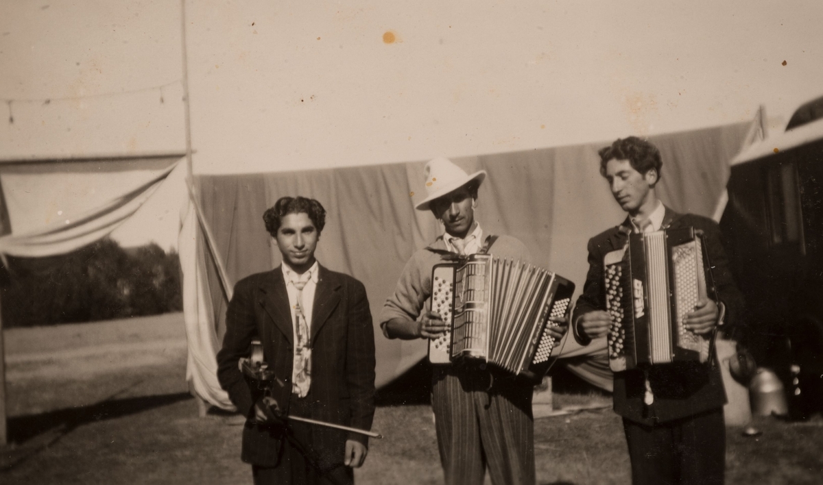 Tre romska män med instrument i ett läger i Sandviken, juni 1947. En av dem håller en fiol och de andra två dragspel. Dukarna som syns i bakgrunden skärmar av lägret och är till för att hålla obehöriga utanför.
