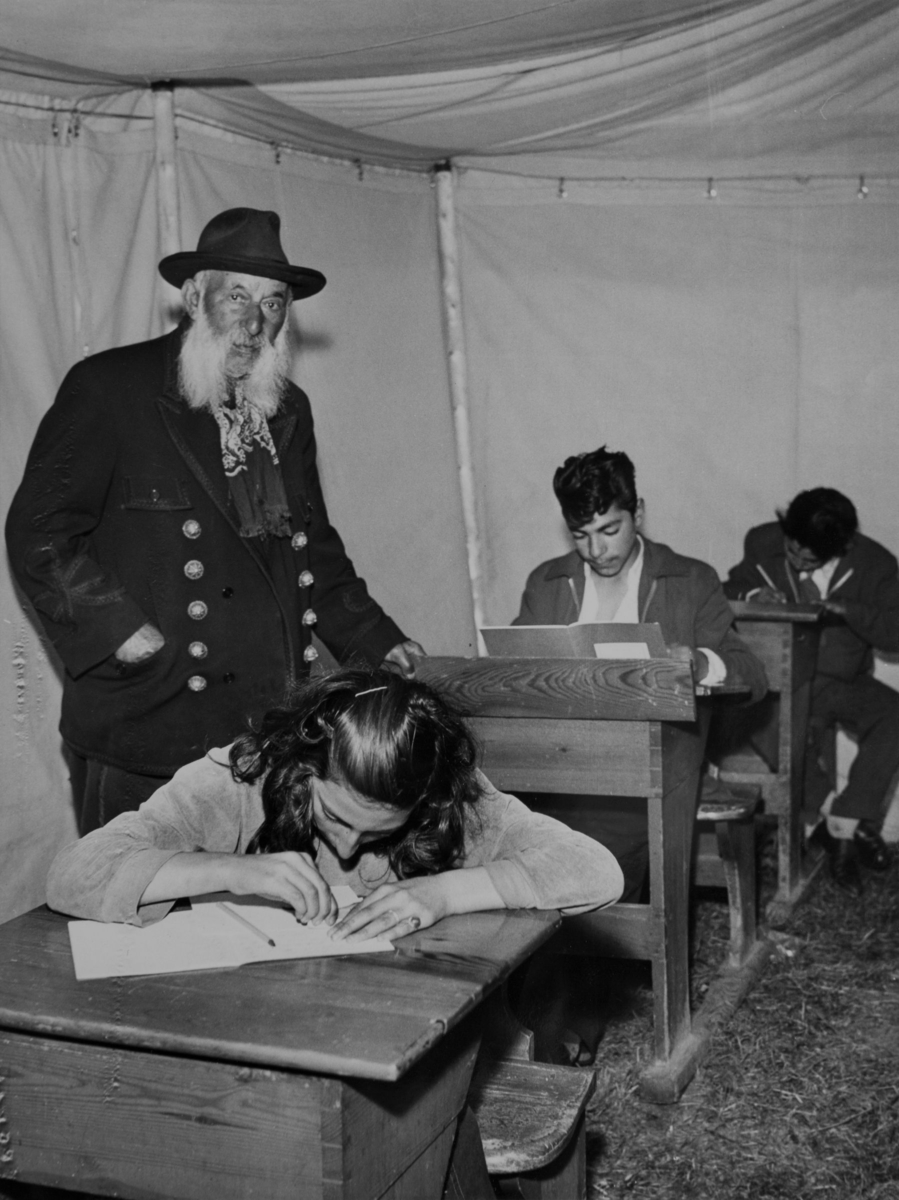 Tre romska elever sitter i sina bänkar i lektionstältet vid sommarskola 1957. En äldre romsk man övervakar dem. Han bär en kavaj med stora knappar av metall.