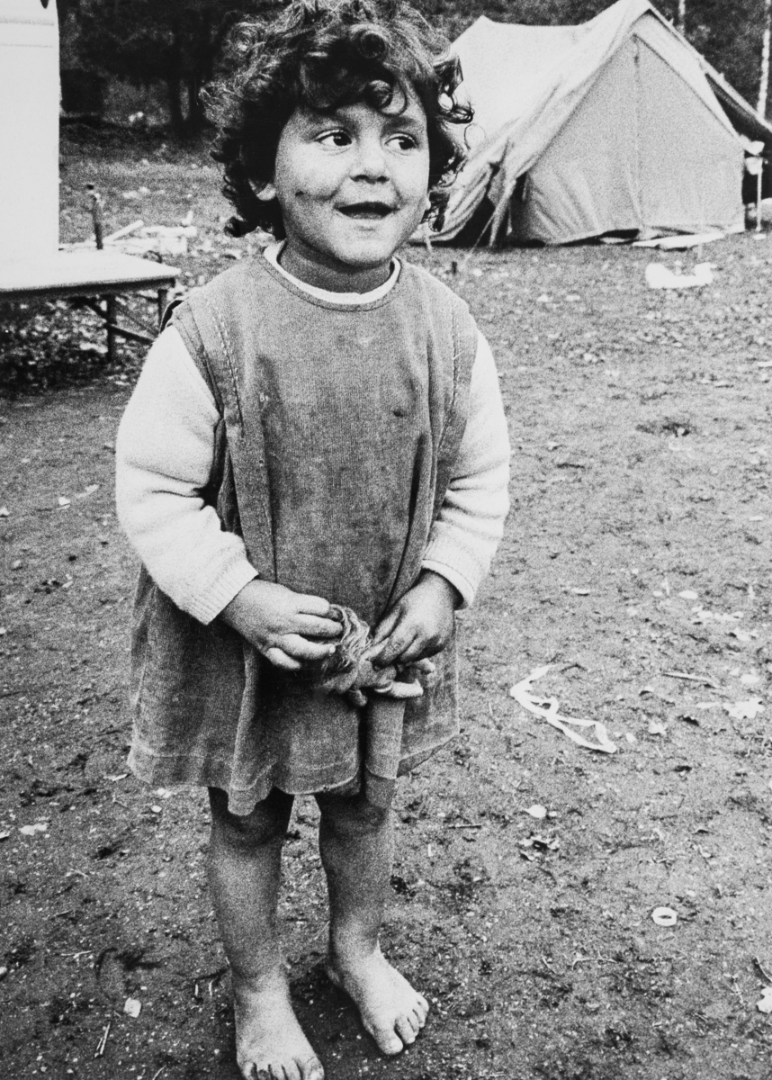 En flicka står barfota med en docka i händerna. I bakgrunden syns tält i det romska läger flickan bor.