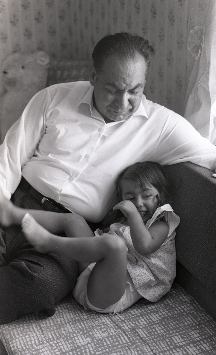 En man sitter i sin soffa med sin dotter intill sig. Bilden är tagen i samband med ett nyhetsreportage för Skånska Dagbladet. Efter att de svenska romerna i Sverige erkändes som medborgare år 1952 uppstod debatt kring gruppens svåra levnadsförhållanden.