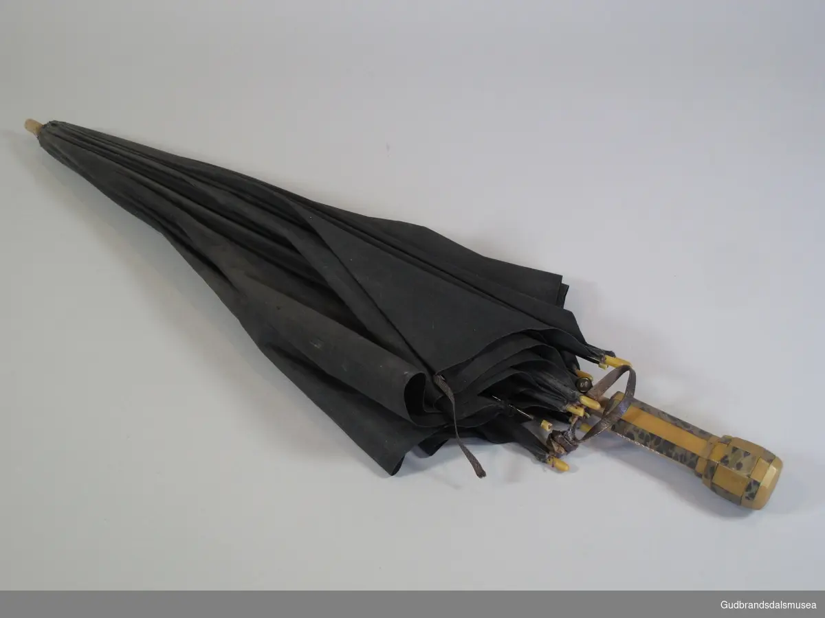 Liten dameparaply fra 1930-50-tallet. Skjerm i svart stoff. Ottekantet håndtak i tre belagt med plastlameller. Spilere i metall. Stang i tre. Liten lærstropp for omvikling av paraply i sammenslått tilstand.